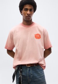 Άνδρας που στέκεται με τα χέρια πίσω από την πλάτη φορώντας ροζ μπλούζα "scotch & soda", μπλε τζιν, καφέ ζώνη και μαύρο μαντήλι με σχέδια που κρέμεται από την θηλιά της ζώνης.