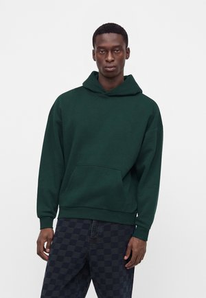 Homme portant un sweat à capuche vert foncé avec poche avant et pantalon sombre à carreaux, debout devant un fond blanc uni.