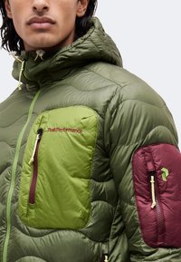 Homme portant une veste matelassée verte avec capuche, avec une poche poitrine verte et une poche de manche bordeaux avec des fermetures éclair.