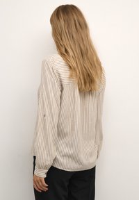 Helle beige Langarmbluse mit vertikalen weißen Streifen, abgerundetem Saum und Knopfdetails an den Ärmeln. Weicher Stoff mit lockerer Passform.