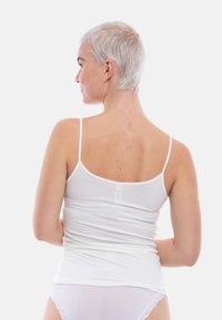 Donna con capelli biondo platino corti, che indossa un top bianco e biancheria intima, vista di spalle su uno sfondo neutro.