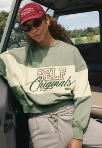 Vrouw in een rode pet, met donkere zonnebril, een groen en crèmekleurig sweatshirt met "SELF Originals 2023" en grijze joggingbroek, zittend in een voertuig.