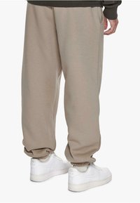 Beige joggings met zijzakken, elastische boorden en een comfortabele pasvorm, gecombineerd met witte sneakers. Het materiaal lijkt zacht en textuur te hebben.