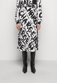 Abito in bianco e nero con motivi testuali astratti, dotato di maniche lunghe e una vestibilità ampia, abbinato a stivali neri alti fino al ginocchio di texture.