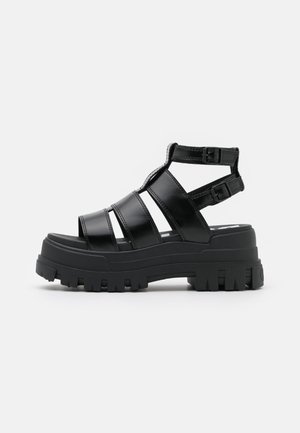 Sort chunky platformsandal med tykke stropper og to justerbare ankelspænder på en ensfarvet lys baggrund.