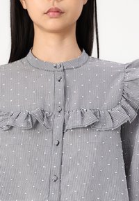 Svart och vit mönstrad blus med rynkad ok, rund krage och knäppning, med ett texturerat rutmönster och prickar.