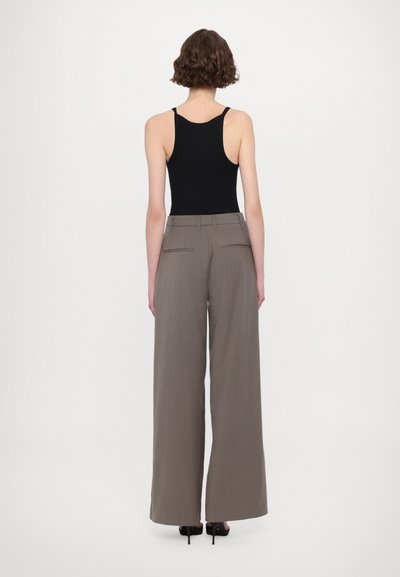 Schwarzes ärmelloses Top kombiniert mit weiten, hochgeschnittenen beigen Hosen mit hinteren Taschen und dezenten Nähdetails. Schwarze Absätze.