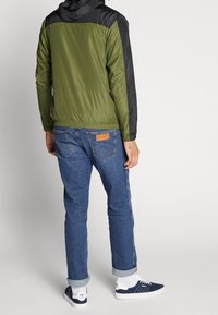 Grøn og sort hættejakke med elastisk talje, båret med blå denim jeans og marineblå sneakers. Har et struktureret stof og logo patch.