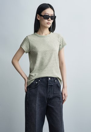 Kvinde med glat sort hår iført sorte solbriller, en lysegrøn stribet t-shirt og løstsiddende sorte jeans mod en grå baggrund.