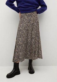 Donna che indossa una lunga gonna floreale fluida, stivaletti neri alla caviglia e un maglione blu scuro con le mani sui fianchi, su uno sfondo neutro.