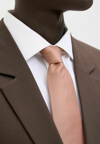 Jachetă de costum maro cu o cămașă albă și o cravată roz deschis, având texturi netede și o croială adaptată, punând accent pe guler și nodul cravatei.