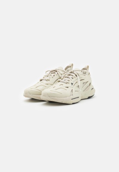 Light beige athletic shoes with a textured upper, mesh panels, contrasting black sole, and lace-up design featuring well-defined cutouts.