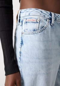 Lyseblå denimjeans har høy midje, frynset kant og en rød Calvin Klein-etikett på baklommen. Stoffet har en litt falmet tekstur.