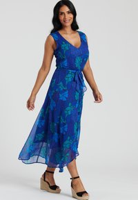 Blå blommig klänning med V-ringning, ärmlös design, knytmidja och genomskinlig överdel, med nyanser av blått och grönt. Bärs med svarta kilklackar.