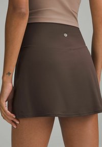 Mujer vestida con una falda deportiva marrón con el logo de Lululemon en la cinturilla y una parte de arriba beige, mostrando un pequeño tatuaje de abeja en la muñeca izquierda.
