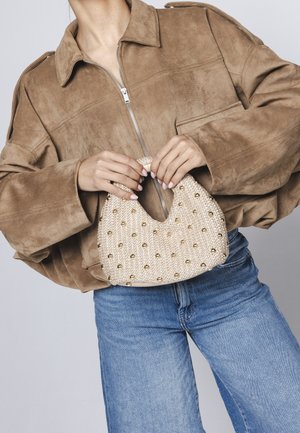 Άτομο με καφέ σακάκι από σουέντ και μπλε τζιν κρατάει μπεζ πλεκτή τσάντα με χρυσά τρουκς μπροστά σε γκρι φόντο.