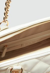 Sac à main matelassé crème avec quincaillerie dorée. Il présente une poche zippée à l'intérieur et un motif de logo sur la doublure. Chaîne de bandoulière visible.