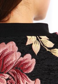 Černá vyšívaná bunda s velkým růžovým květinovým motivem, s texturovanou látkou a žebrovaným černým límcem. Viditelné detailní květinové akcenty.