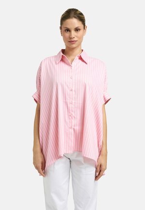 Woman wearing loose pink short-sleeve button-up shirt with white vertical stripes and white pants, standing against a plain background.