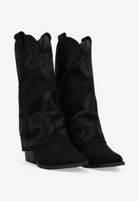 Botas de vaquero de ante negro con tachuelas decorativas en tono plateado, punteras puntiagudas y un tacón robusto. Presenta un detalle de diseño curvado en el cañón.