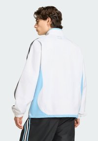 Νέος άνδρας με κοντά καστανά μαλλιά, που φοράει λευκό και ανοιχτό μπλε μπουφάν και μαύρο παντελόνι με μαύρες ρίγες, κοιτάζει προς τα πίσω με το κεφάλι στραμμένο πλαγίως.