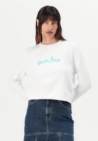 Baltas trumpas sportinis džemperis su turkio spalvos "Guess Jeans" logotipu, ilgomis rankovėmis, apvaliu iškirpimu, derinamas su tamsiai mėlynu džinsiniu sijonu. Lygus audinys.