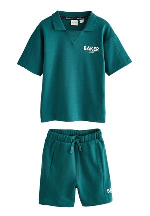 Mladý chlapec v tyrkysovej mikine a kraťasoch Baker, biele ponožky Baker a biele tenisky stojí na svetlo šedej podlahe.