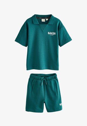 Junger Junge trägt ein türkisfarbenes Baker-Sweatshirt und Shorts, weiße Baker-Socken und weiße Turnschuhe und steht auf einem hellgrauen Boden.