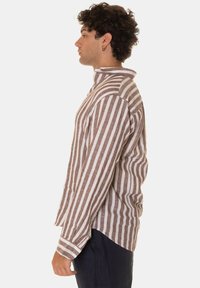 Camicia in lino a righe beige e bianco, caratterizzata da un colletto alto, maniche lunghe e un fit rilassato. Dettaglio delle cuciture laterali e dei polsini visibile.