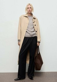 Manteau long beige, pull gris à motifs, pantalon noir à jambes larges et sac fourre-tout en cuir suédé marron foncé. Le modèle se tient avec les cheveux courts.