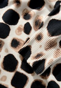 Pletena tkanina v kremni barvi, ki ima design leoparda z velikimi črnimi in bež madeži. Na robovih je vidna rebrasta tekstura.