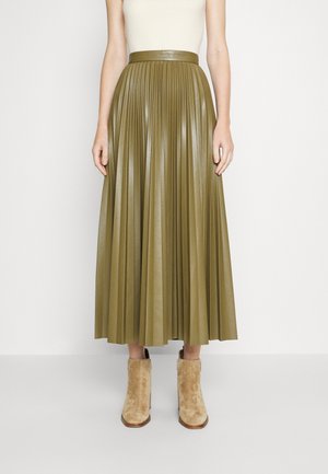 Vrouw met een olijfgroene geplooide midi-rok, wit mouwloos topje en beige suède enkellaarsjes, staand tegen een eenvoudige achtergrond.