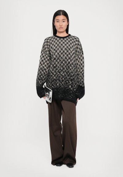 Black and cream patterned sweater with oversized fit and long sleeves, paired with brown wide-leg trousers and holding a clear clutch.