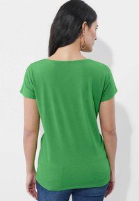 Grünes, kurzärmeliges Oberteil mit legerem Schnitt und rundem Halsausschnitt. Glatte Textur, einfarbig und leicht abgerundeter Saum, kombiniert mit blauer Jeans.