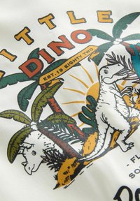 Graphic t-shirt featuring colorful dinosaur and palm tree illustrations, with text "LITTLE DINO" and "EST. 1982" on a white background.