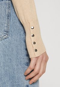 Manche de pull beige tricoté avec des accents de boutons argentés, associée à un tissu en denim bleu clair présentant une texture délavée et des coutures visibles.