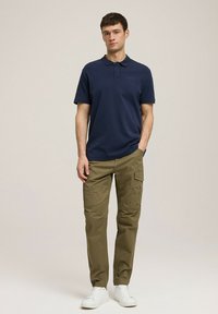 Granatowa koszulka polo z krótkim rękawem, ciemnozielone spodnie cargo i białe sneakersy, stojąca na neutralnym tle.