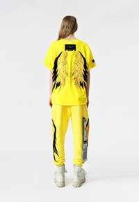 Camiseta amarilla de gran tamaño con gráficos de alas en negro y dorado en la parte trasera, combinada con pantalones de chándal a juego y botas blancas.