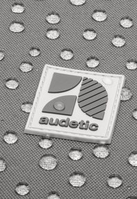 Šedá textúrovaná tkanina s vodnými kvapkami, so bielou obdĺžnikovou nášivkou s geometrickými tvarmi a slovom "audetic" v šedej farbe.