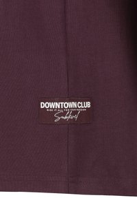 Burgunderrotes Baumwoll-T-Shirt mit einem rechteckigen Etikett, auf dem "DOWNTOWN CLUB" und "Sublevel" in Weiß auf einem dunkelbraunen Hintergrund steht.