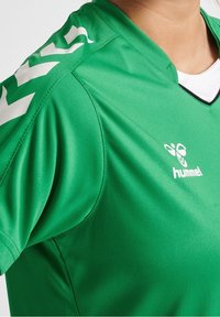 Camiseta deportiva verde hecha de una tela suave y liviana. Cuenta con un cuello en V y un logo blanco con acentos en los hombros.