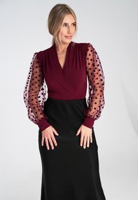 Bordowa sukienka o kroju kopertowym, z przezroczystymi, kropkowanymi bufkami oraz gładkim czarnym materiałem poniżej talii, tworząca elegancką sylwetkę.