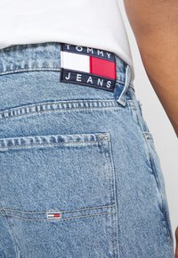 Ljusblå denimjeans med en Tommy Jeans-märke i röd, vit och blå. Nära bild av bakfickan och midjebandets detaljer.