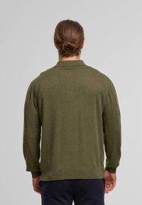 Man with brown hair tied in a bun wearing an olive green long-sleeve sweater and dark pants, standing with back facing the camera.