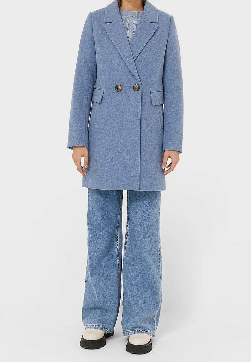 Personne portant un manteau bleu clair à double boutonnage, un jean à jambes larges bleu et des chaussures blanches à semelles noires, debout devant un fond uni.