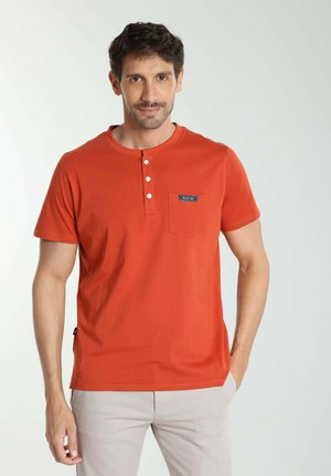 Homme portant une chemise henley orange à manches courtes avec poche avant et un pantalon beige, debout devant un fond clair uni, regardant vers l'avant.