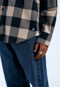 Camisa de franela a cuadros en azul marino y beige, abotonada con un logo de parche. Combinada con pantalones de mezclilla azul oscuro y un anillo de plata en la mano.