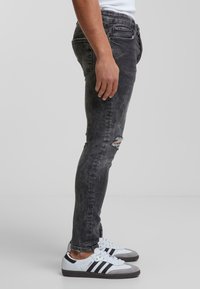 Feszes farmernadrág elhalványult fekete denimből, térdnél szakadt részlettel, fehér sportcipővel párosítva, szürke kiegészítőkkel és fekete csíkokkal.
