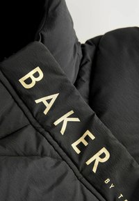 Collier de veste en doudoune noire avec une texture mate, arborant des lettres dorées en relief "BAKER by T" sur le tissu.