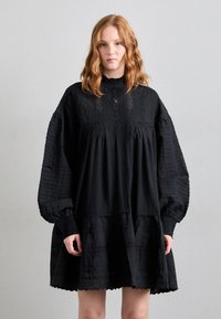 Črna obleka z visokim ovratnikom, ki ima teksturirane pihane rokave, detajle s fizičnimi gumbi ter večplastno krilo z čipkastim robom in naborki.